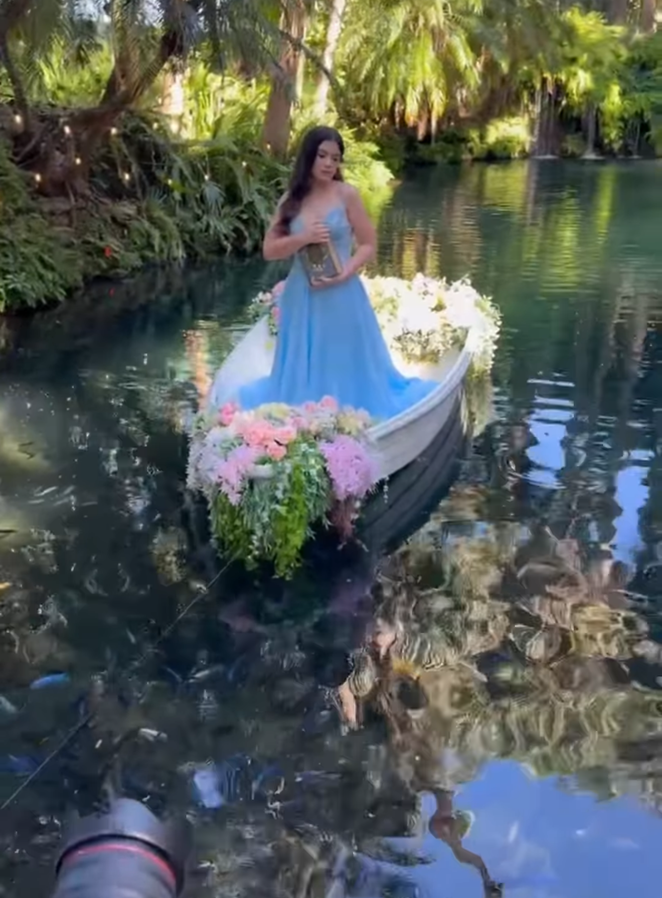 Woman in blue gown standing on flower-adorned boat gliding through a serene tropical waterway.