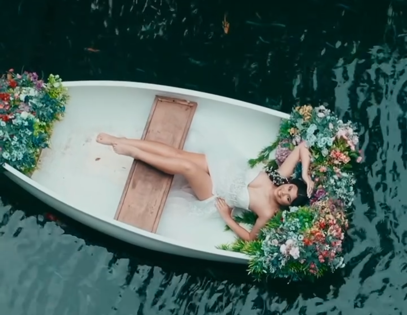 Woman lying in a flower-adorned white rowboat floating on dark water, aerial view.