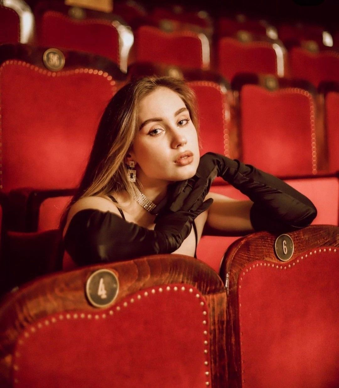 Young woman in black gloves and off-shoulder dress poses elegantly among red velvet theater seats.