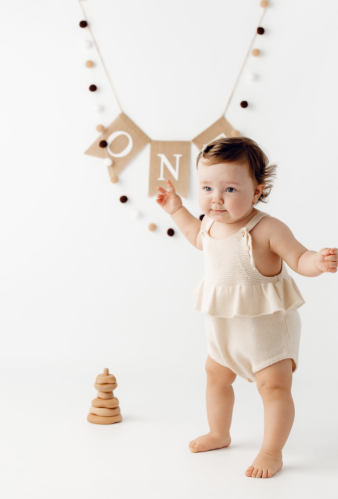 A baby girl in a cream romper stands barefoot near a wooden ONE banner for her first birthday photo session.