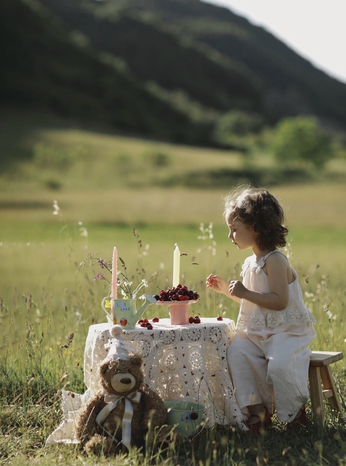 Young girl at a whimsical outdoor tea party with cherries, candles, and a teddy bear in a lush green meadow.