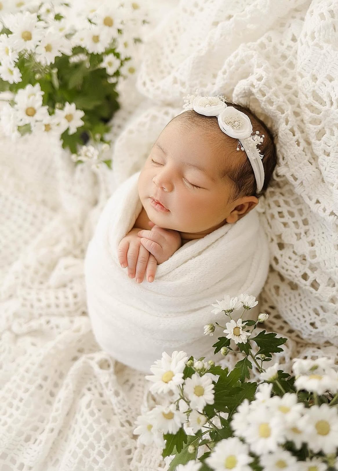 Newborn baby swaddled in white wrap with floral headband, sleeping peacefully among white daisy flowers on lace blanket.