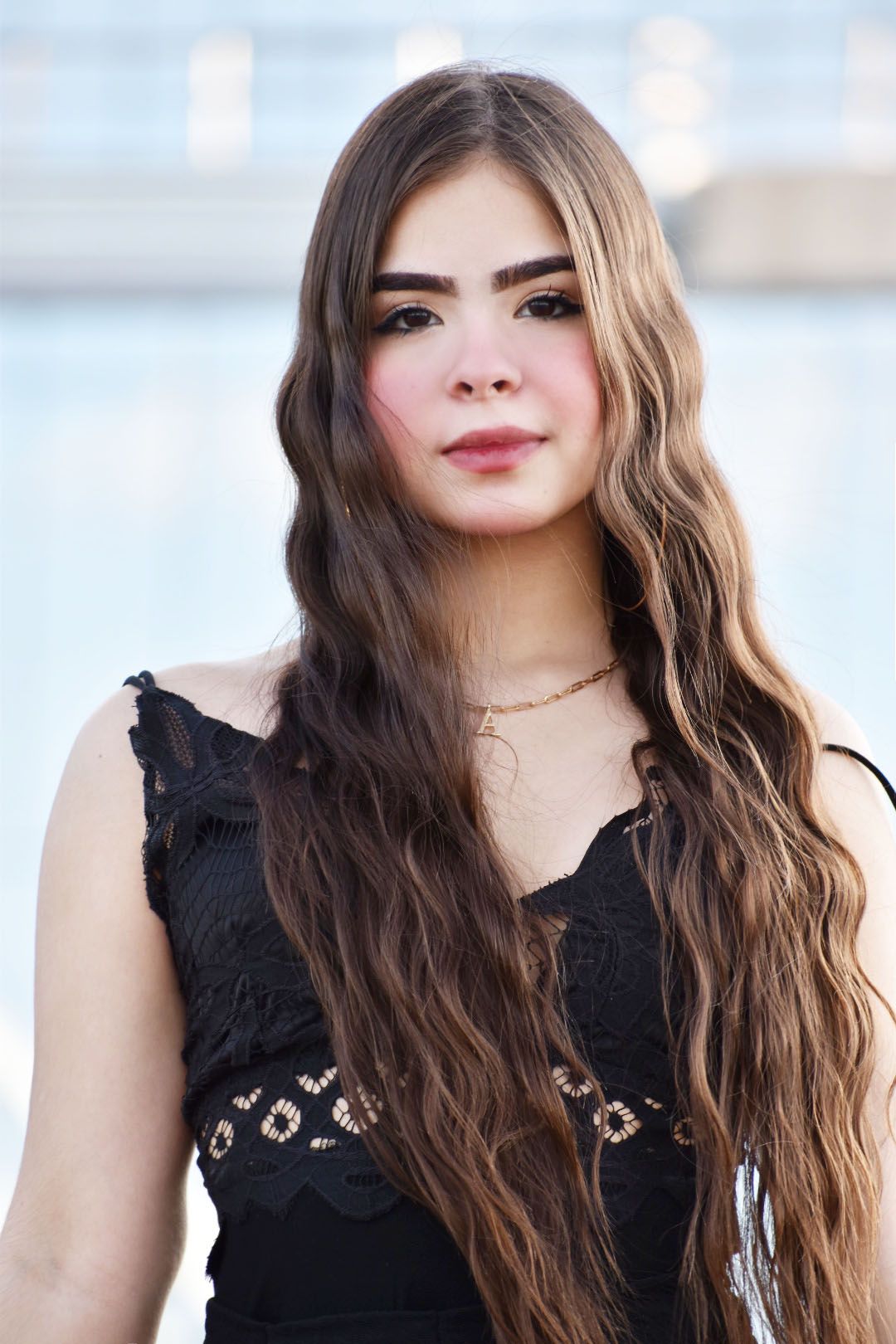Portrait in black lace top with wavy brown hair and delicate necklace against blurred background.