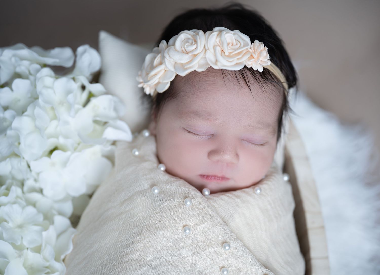 Delicate newborn portrait with white rose headband against cream background.