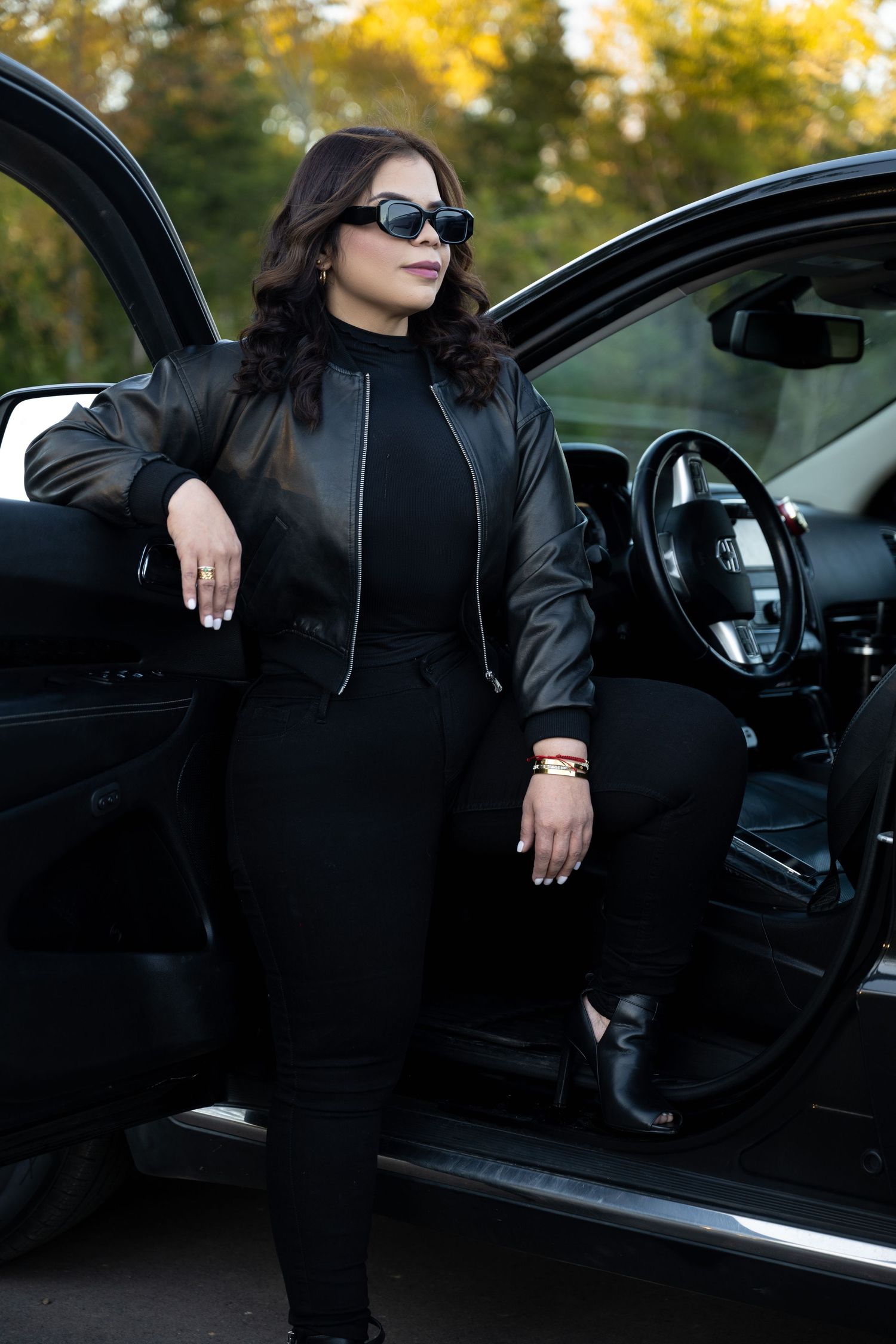 Stylish black outfit and sunglasses pose by luxury car door in autumn setting.