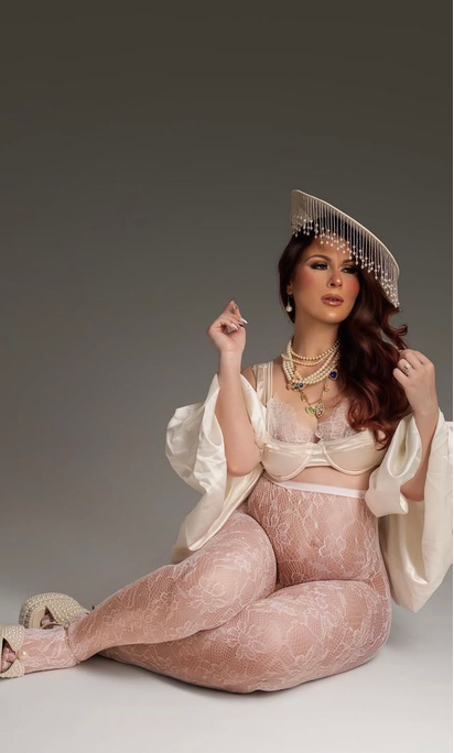 Woman with wavy auburn hair sits in a studio wearing a beaded fringe headpiece, pearl necklaces, pink lace tights, and a cream bralette.