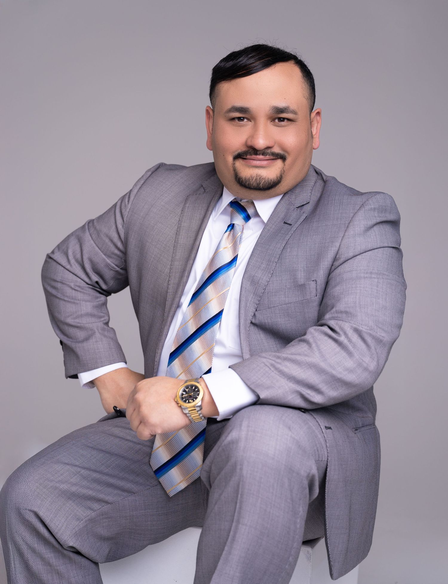 Smiling man with a goatee in a gray suit, striped blue tie and gold wristwatch sits confidently against a light gray studio background.