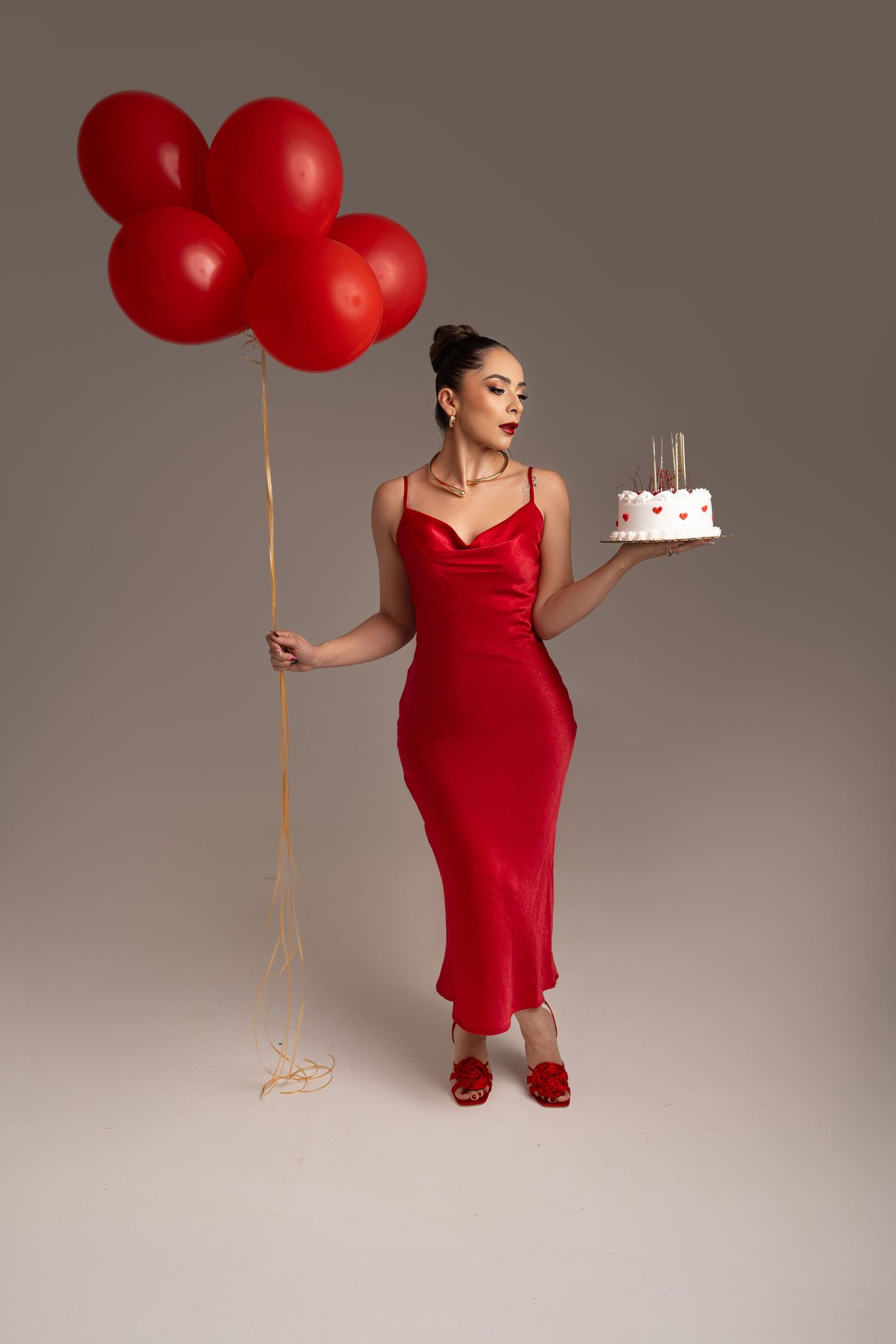 Woman in a red satin slip dress and red heels holds a bunch of red balloons and a small birthday cake with candles against a gray backdrop.