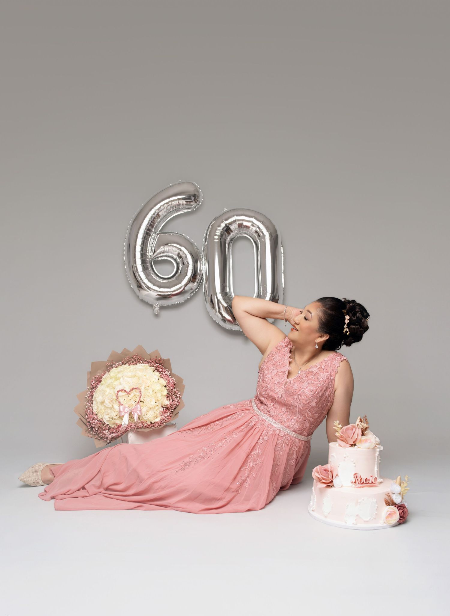 Woman in pink gown sits beside birthday cake and flowers, celebrating 60th birthday with silver balloon numbers.
