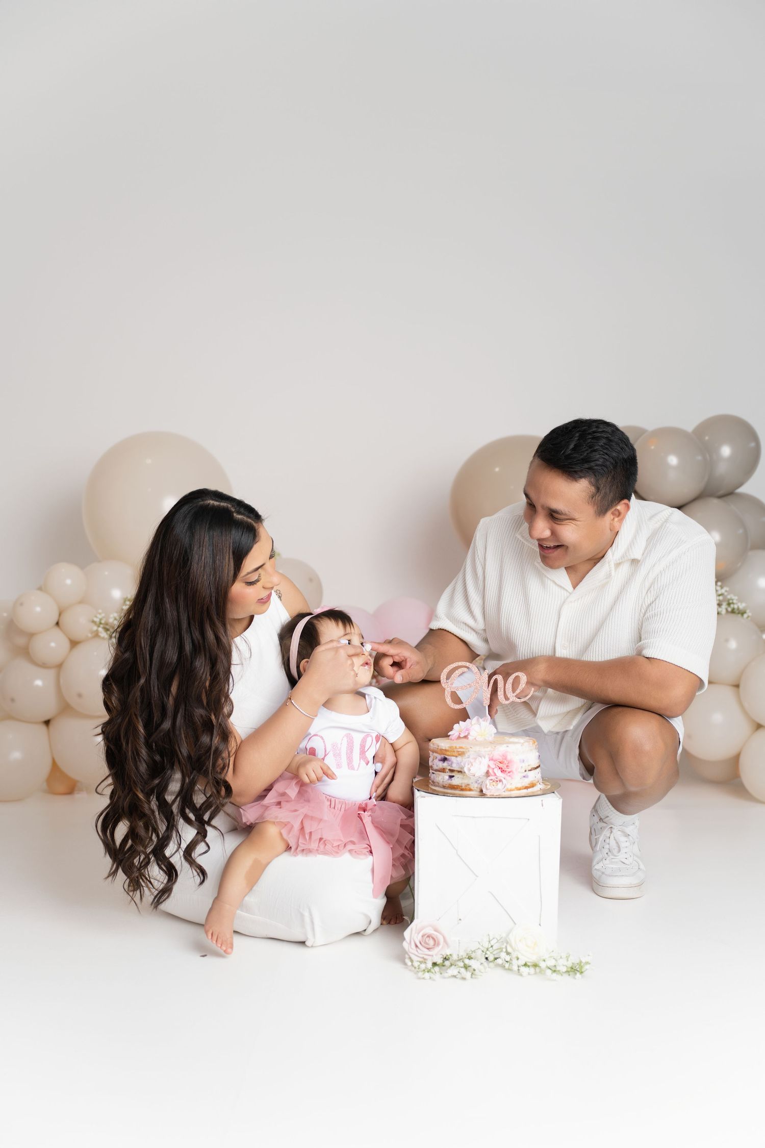 A family celebrates their baby girl's first birthday with a cake smash session against a balloon backdrop.