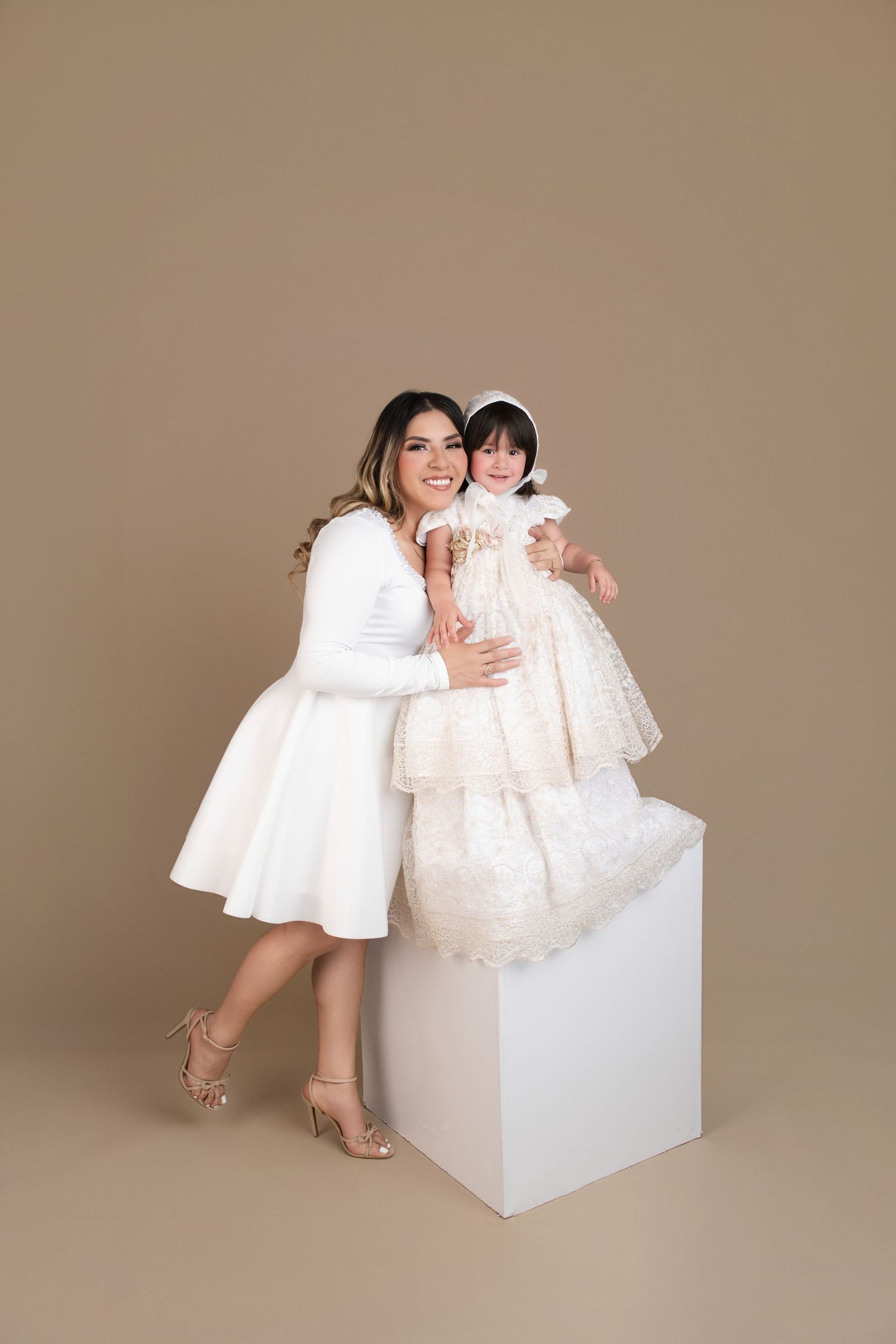 A woman and young girl in matching white dresses pose together in a studio with a beige background.