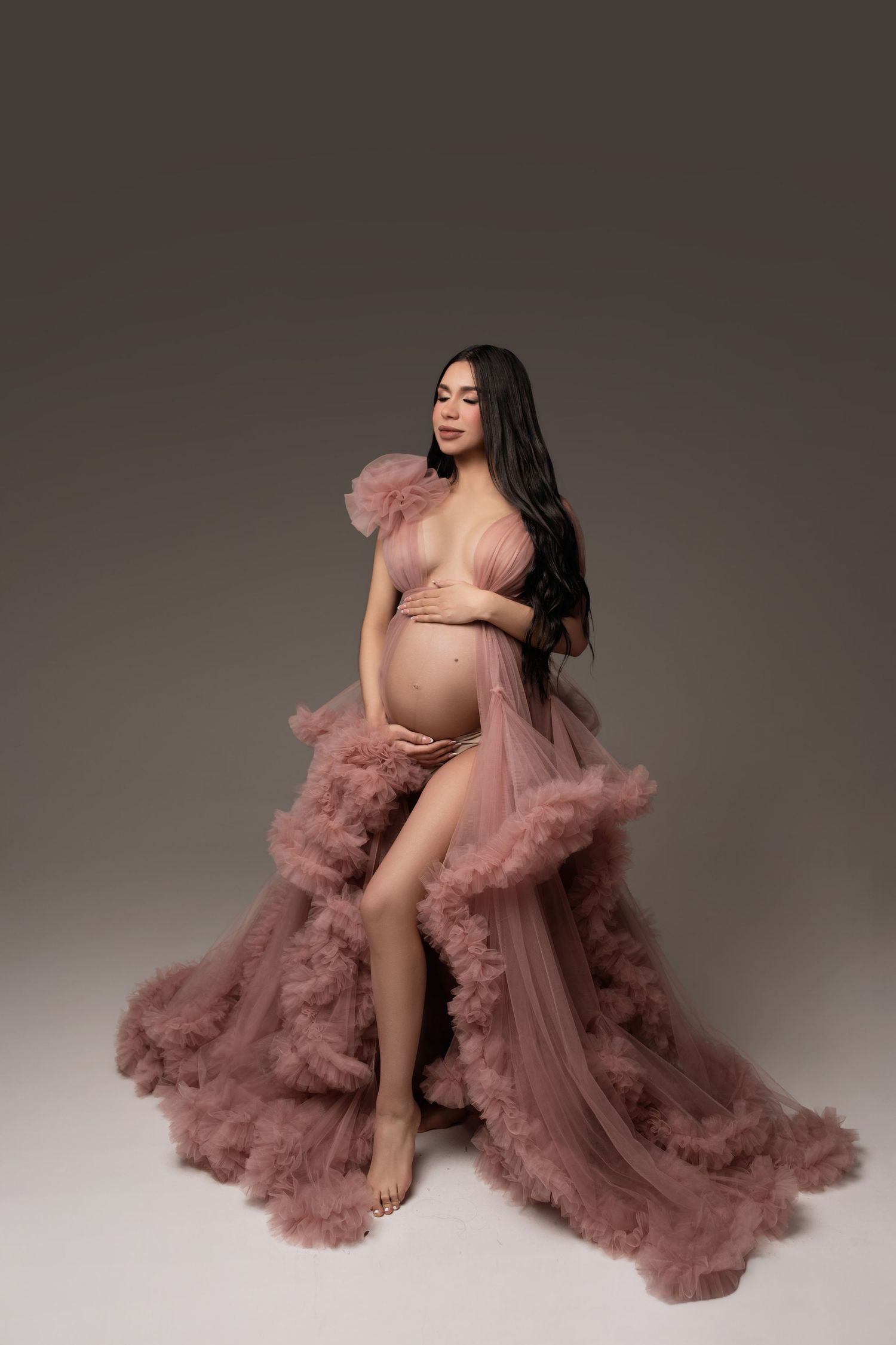 Pregnant woman in dusty rose tulle robe gown posing elegantly in studio maternity photoshoot.