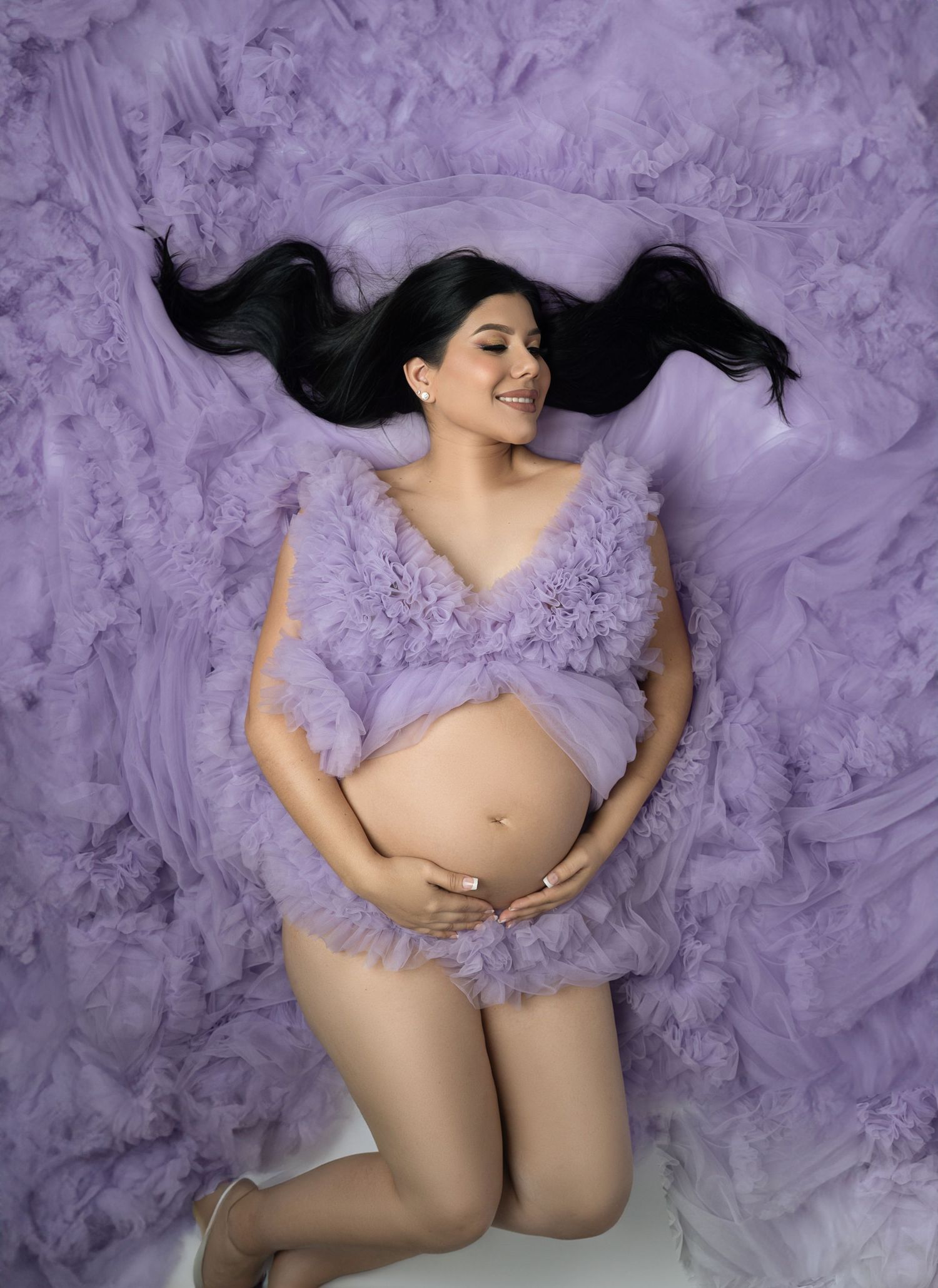 Pregnant woman in lavender tulle robe lying down, cradling baby bump, with long dark hair spread out.