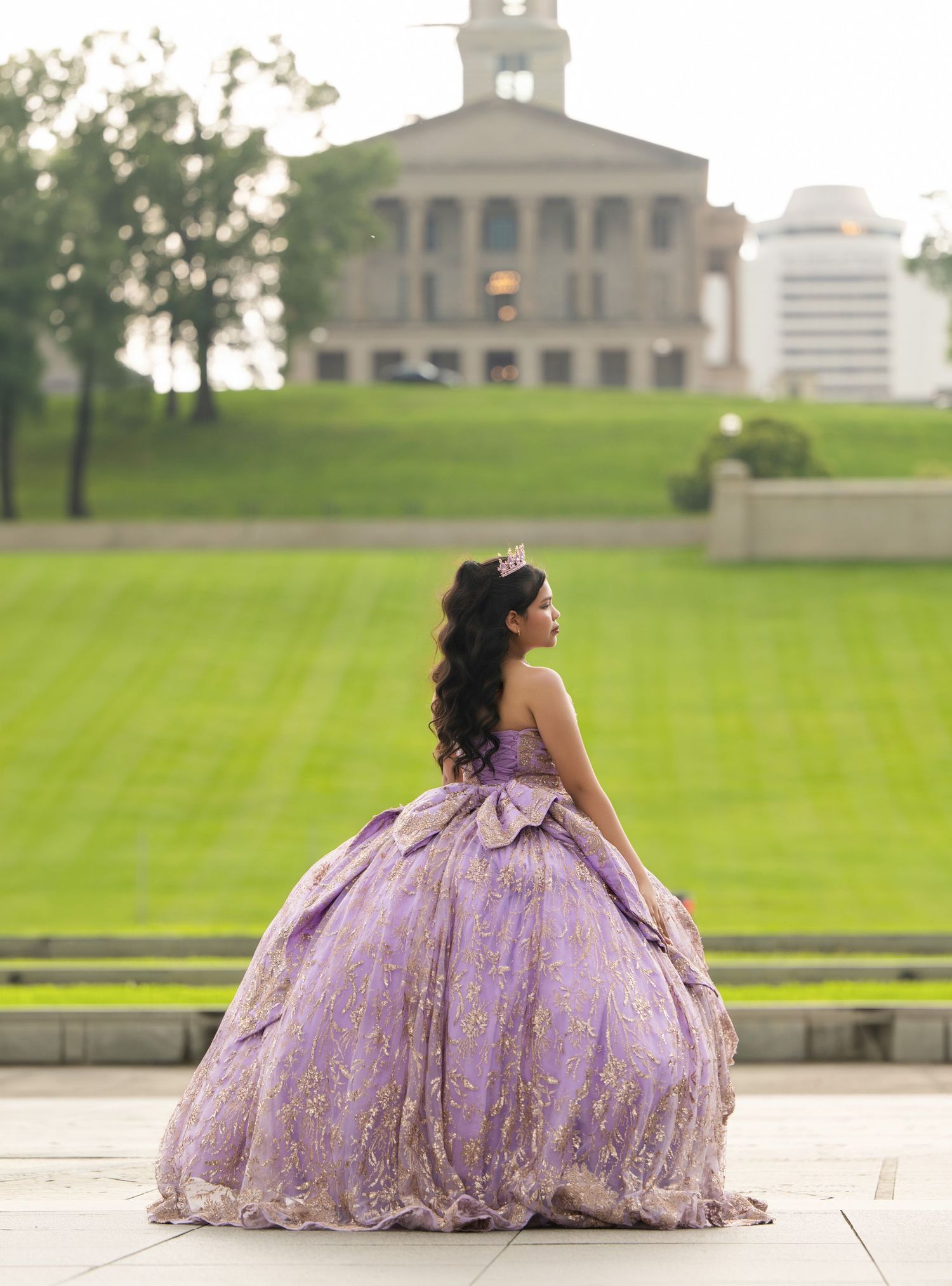 Young woman in lavender quinceañera ball gown with tiara poses on steps before a grand historic building.