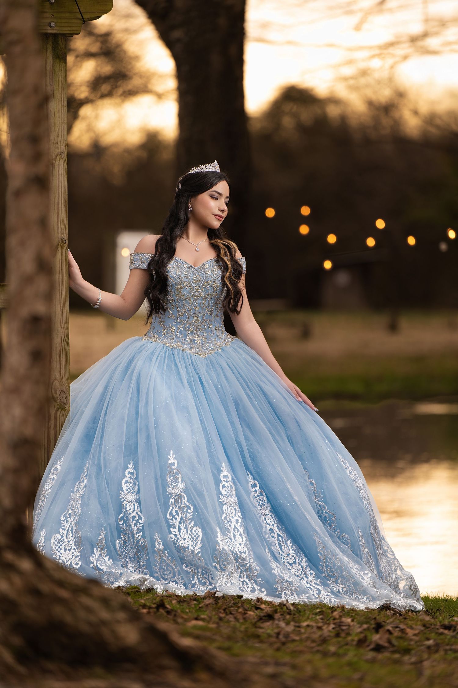 Young woman in a light blue ball gown with lace details and tiara poses outdoors at sunset for quinceañera.