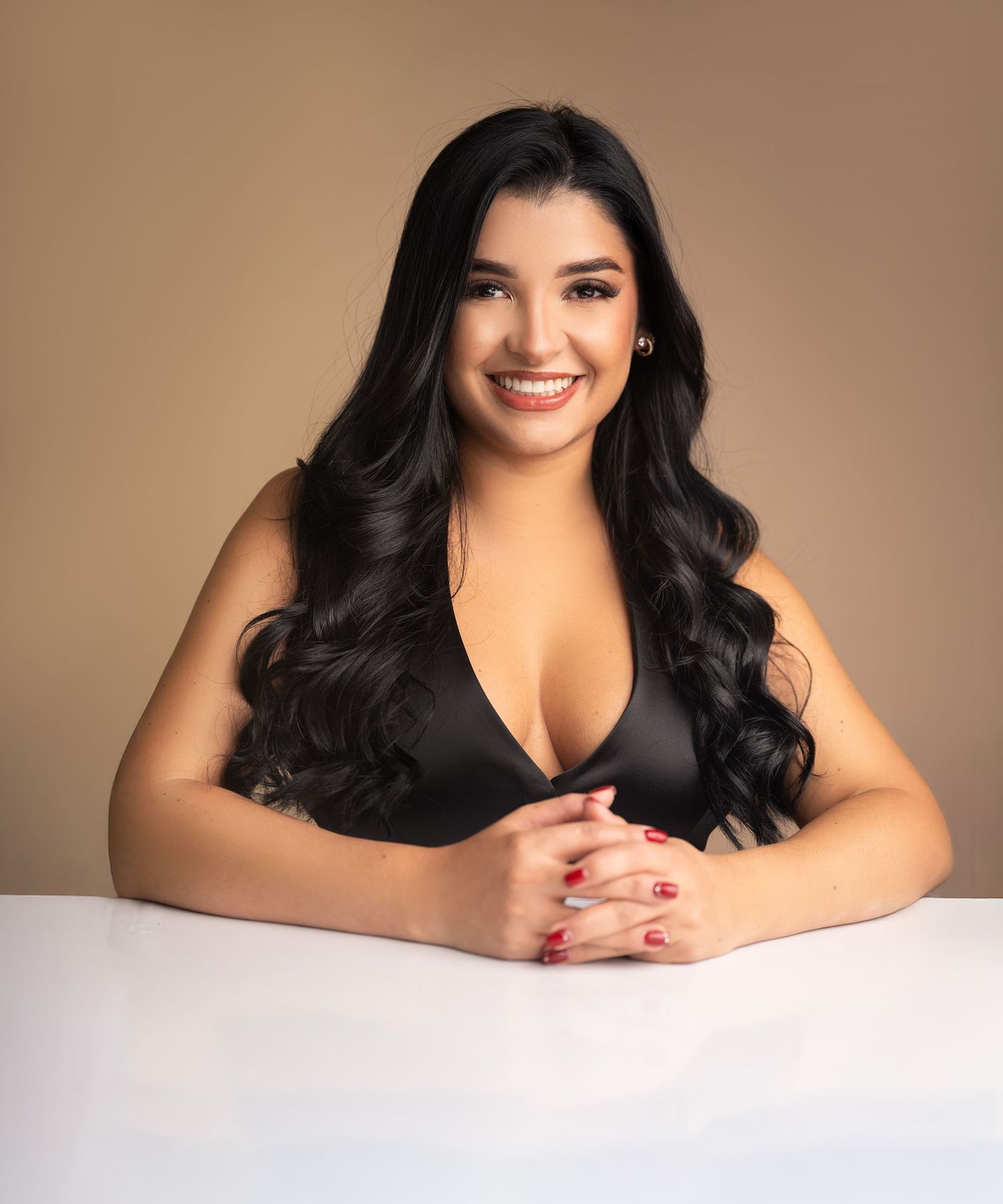 Professional studio portrait of a woman with long dark wavy hair wearing a black top, seated at a white table against brown backdrop.