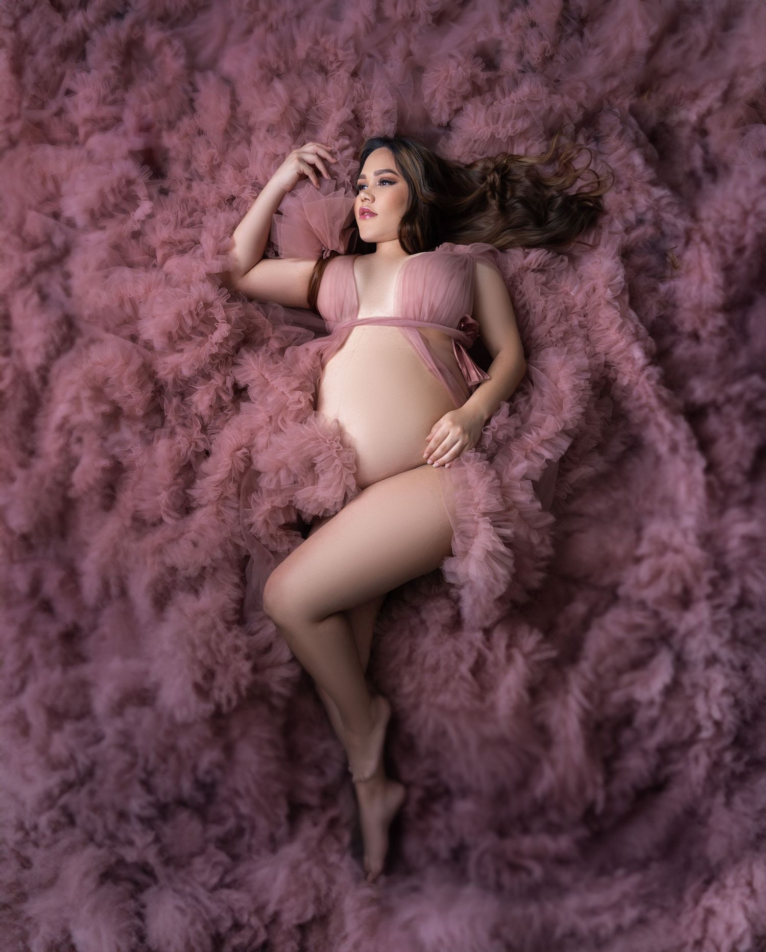 A maternity photo featuring a pregnant woman in pink lying on soft pink cotton candy-like clouds in a dreamy setting.
