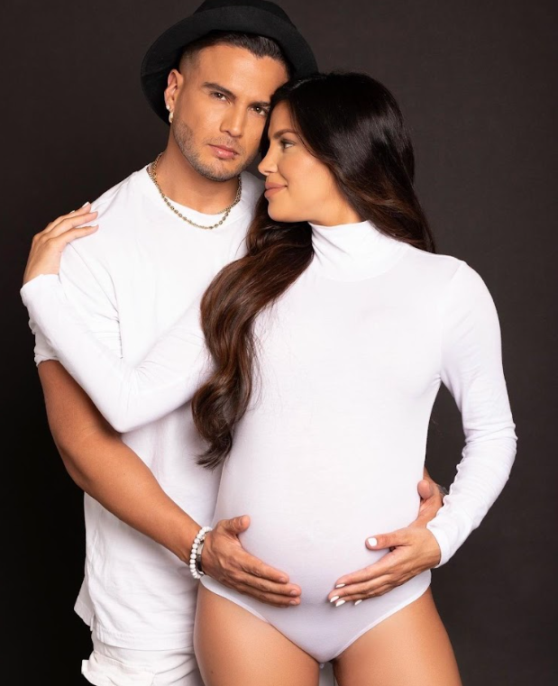 A couple in white outfits embraces intimately during a maternity photo shoot against a dark background.