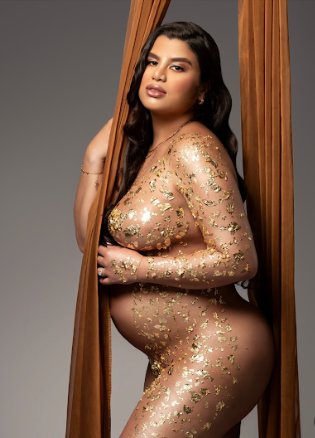 A maternity photoshoot features a figure draped in sheer gold fabric with floral details against a dark backdrop.