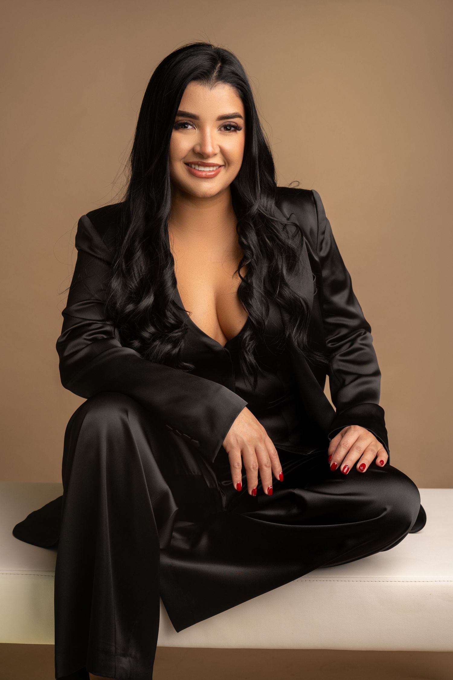 Professional studio portrait of someone in a black satin suit sitting on a white bench.