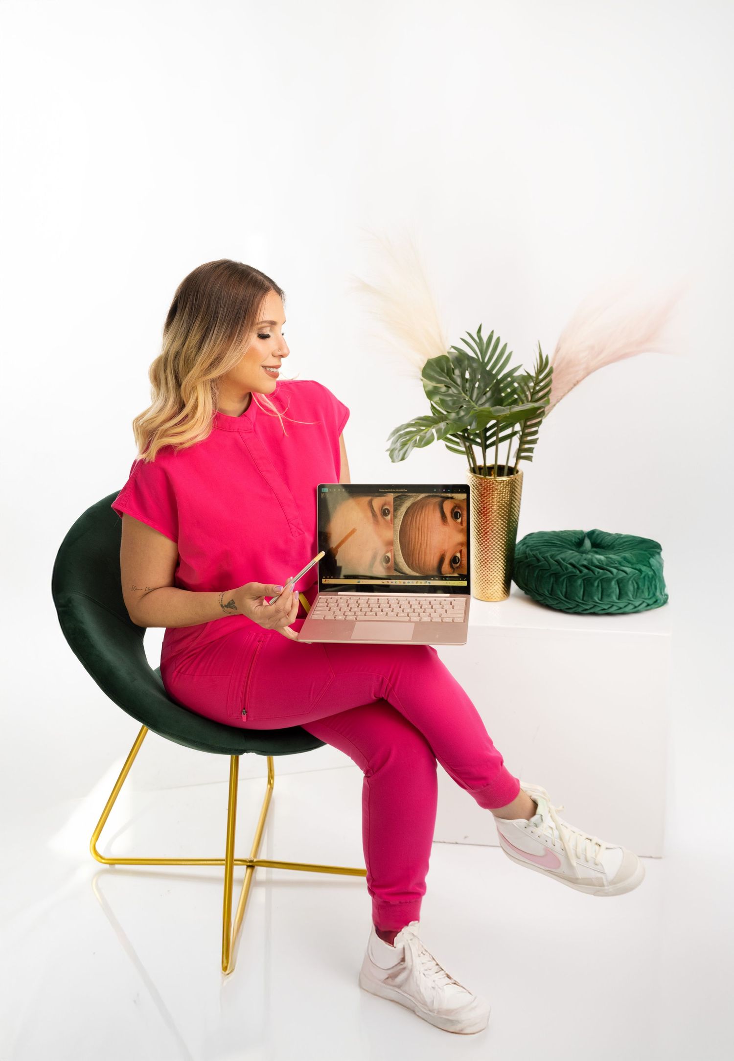 Person in bright pink outfit sits in emerald velvet chair while looking at laptop computer display.