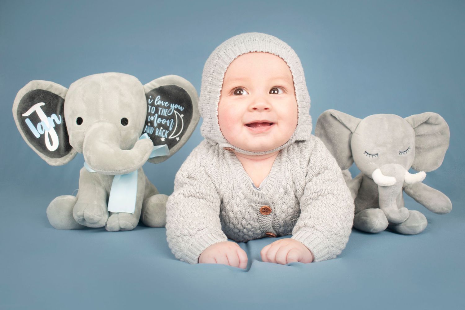 Baby in knit bonnet lying between two plush elephant toys against blue backdrop.
