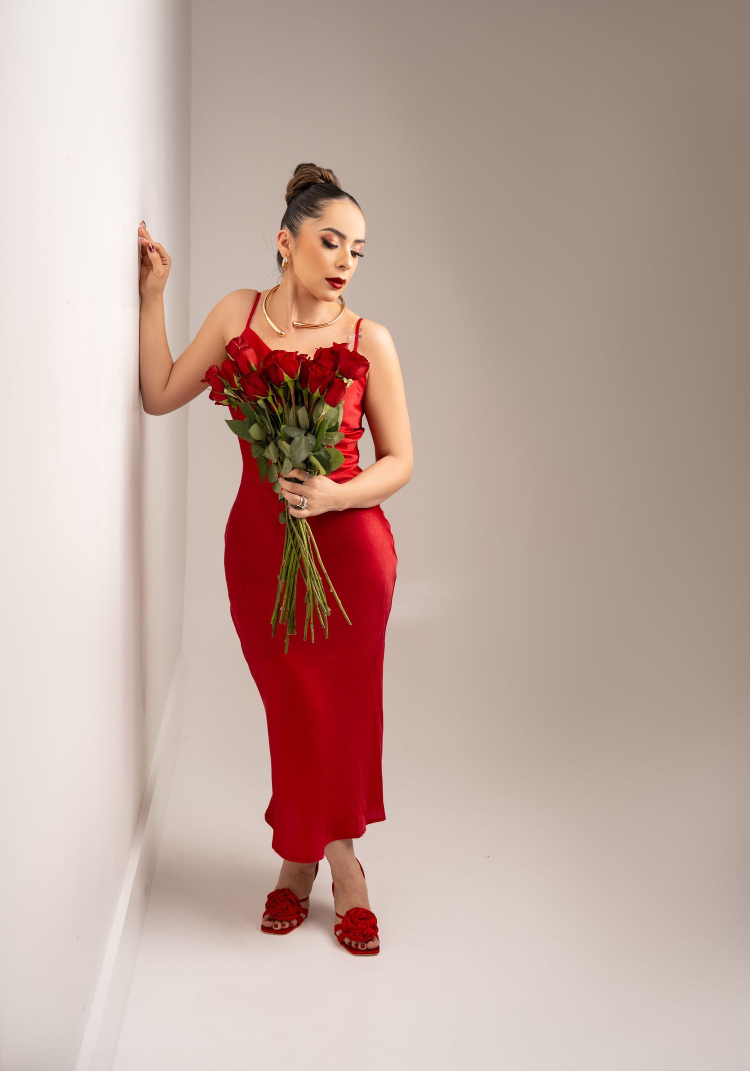 Figure in elegant red slip dress holding fresh roses against neutral background with soft lighting and minimalist styling.