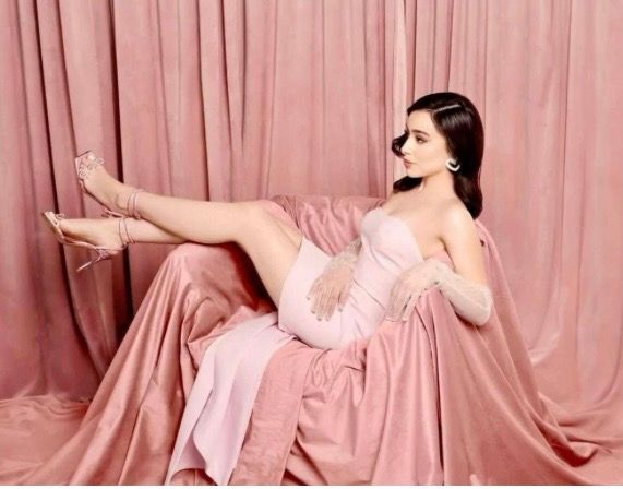 Woman in pink dress reclines gracefully on pink chair against pink curtain backdrop, wearing heels and hair accessory.