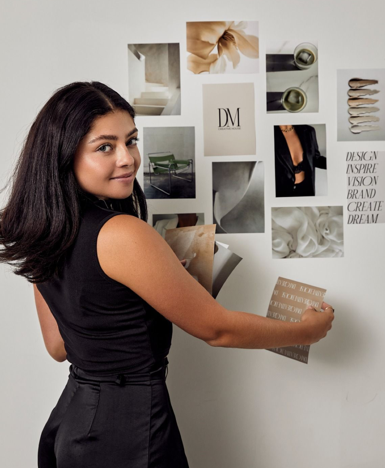 Woman in black dress arranging design inspiration mood board with elegant photos and DM monogram on white wall.