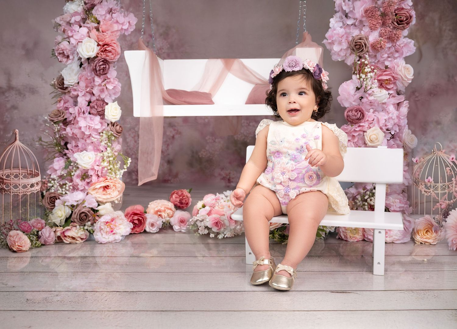 Adorable baby girl in floral romper sitting on white bench surrounded by pink flowers and decorative birdcages.
