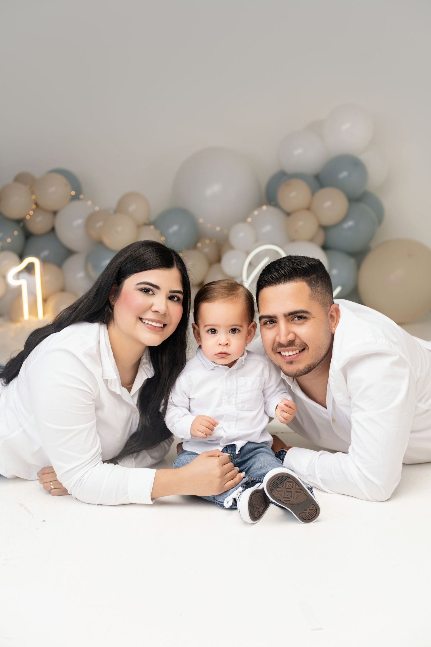 Family of three posing for a first birthday photo with blue and beige balloon decorations and number one backdrop.