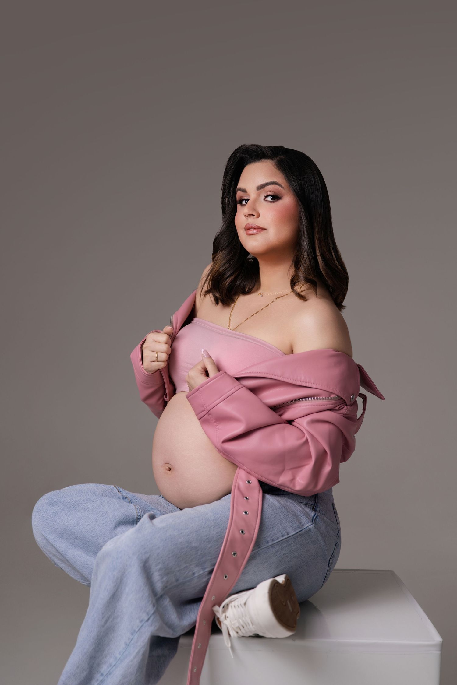Pregnant woman in pink jacket and jeans sitting on white cube, showcasing baby bump in a studio maternity photoshoot.
