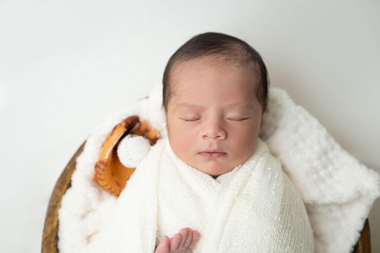 Sleeping newborn wrapped in white swaddle with brown teddy bear prop on white background.