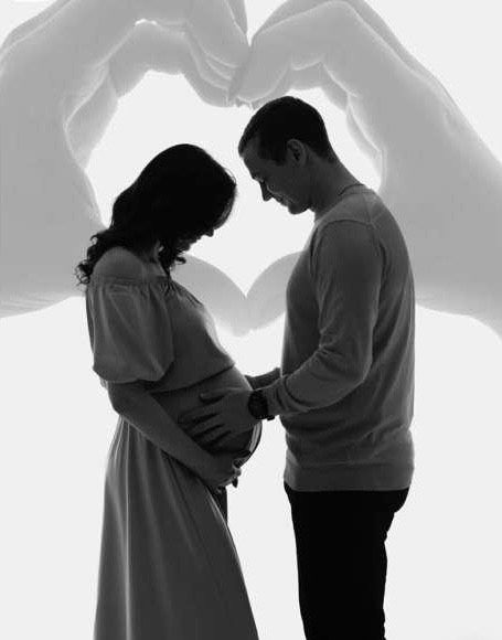 Black and white maternity photo of a couple holding hands over pregnant belly with heart shadow on wall.