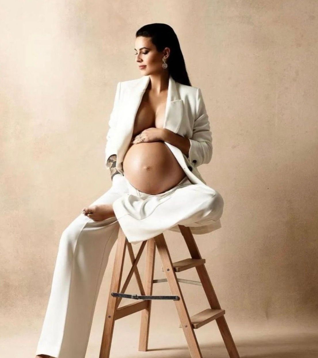 Pregnant woman in white blazer and pants posing on ladder stool, elegant maternity photoshoot.