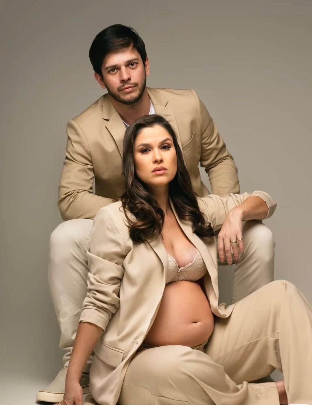 Elegant maternity photo shoot featuring a pregnant woman in beige suit with her partner in a studio setting.