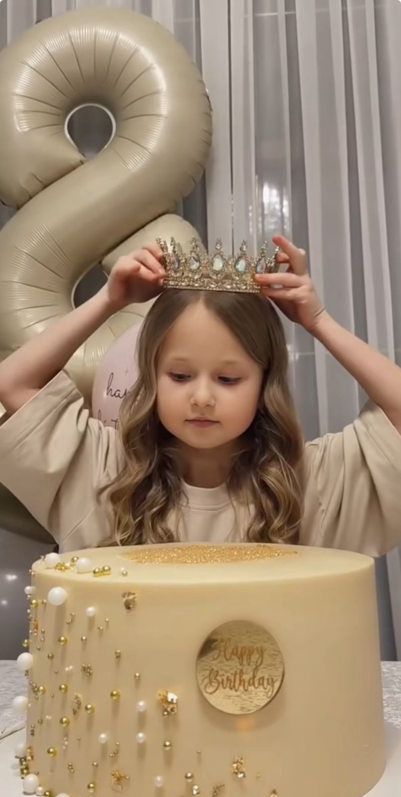 A young girl places a jeweled crown on her head in front of a golden birthday cake and number 8 balloon.