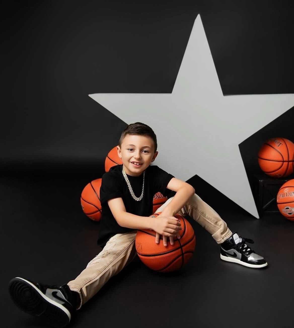 Young boy smiling with basketballs and a star backdrop in a sports-themed portrait photo.