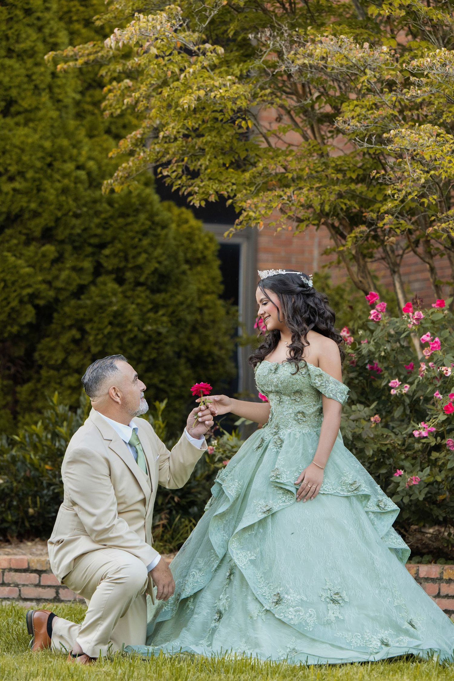 A man kneels presenting a rose to a woman in a sage green quinceañera gown outdoors amid lush greenery.