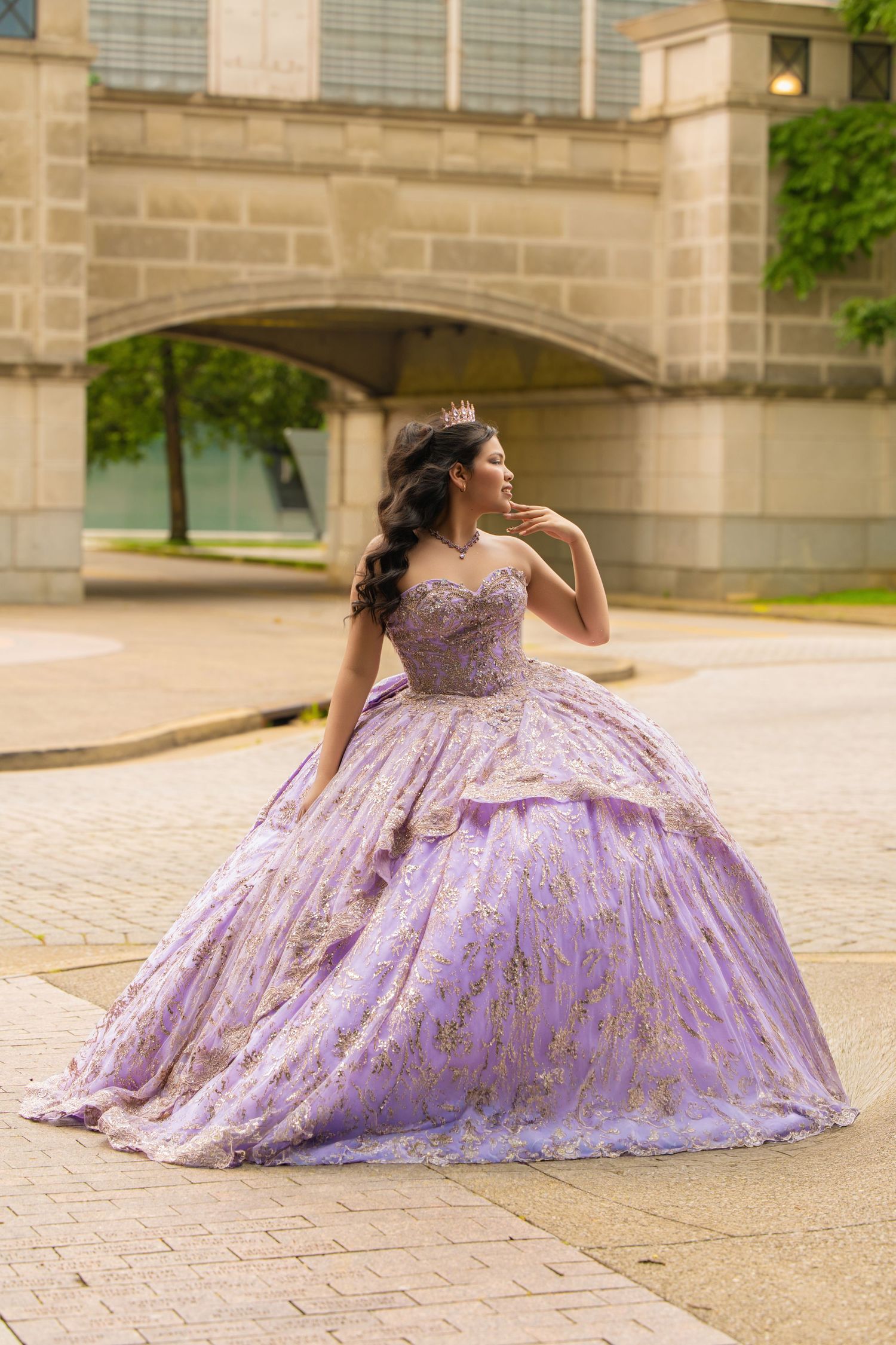 Young woman in lavender quinceañera ball gown with gold details and tiara poses outdoors near stone architecture.