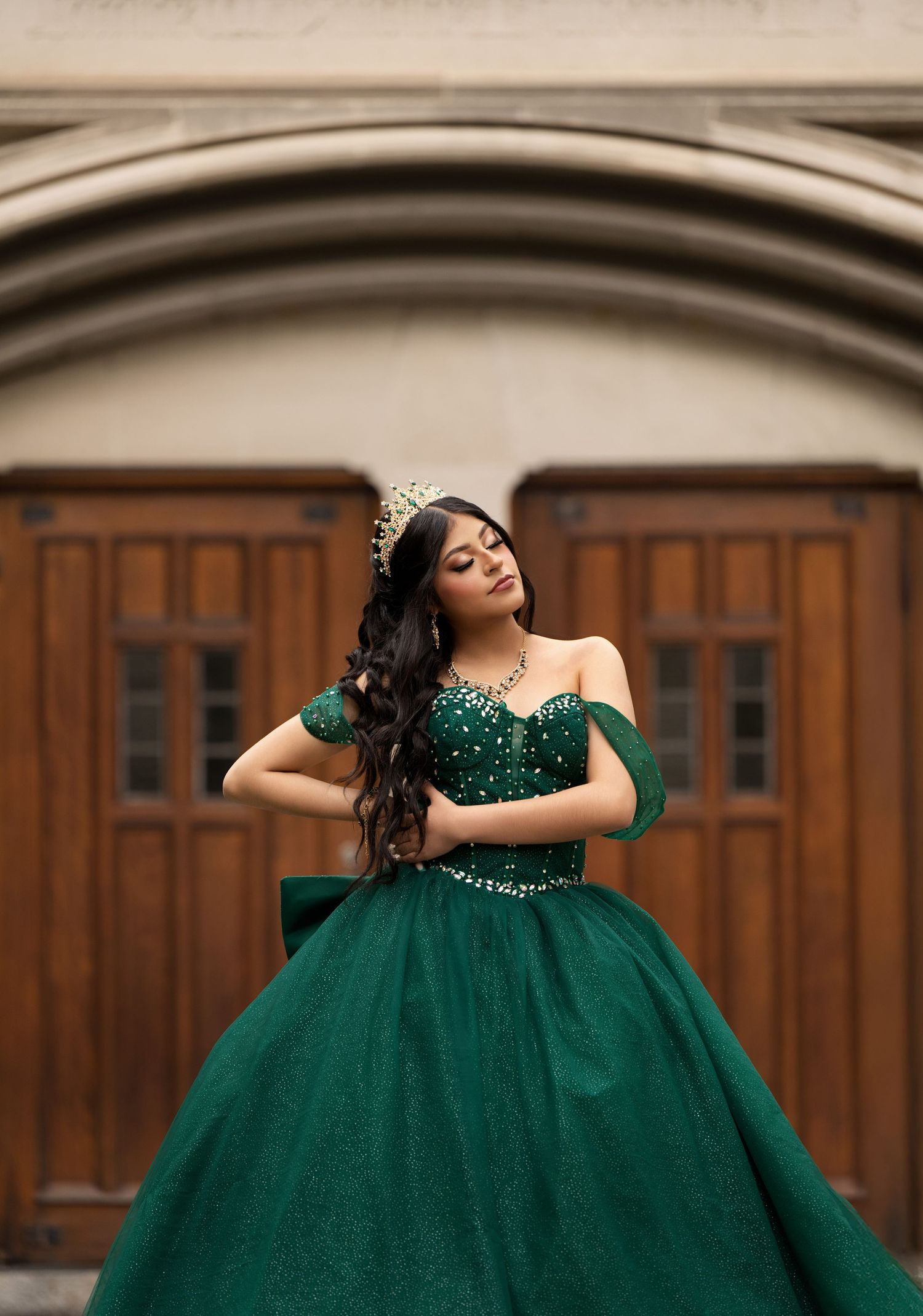 Young woman in emerald green off-shoulder ball gown with crown poses before ornate wooden doors.
