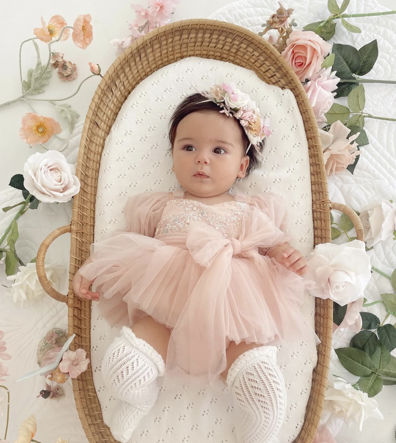 Baby girl in pink tulle dress and floral headband lying in a wicker Moses basket with flowers.