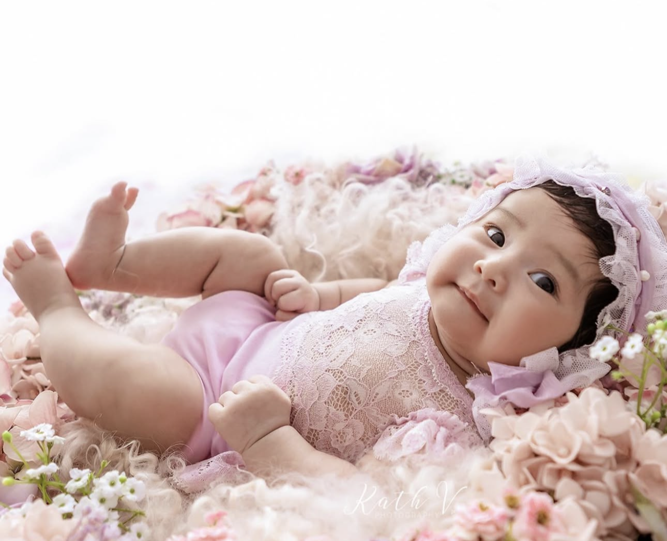 Baby girl in pink lace romper and bonnet lying among soft pink and white flowers in professional photo shoot.