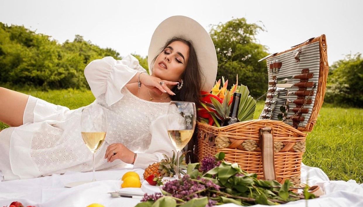 Woman in white dress and hat enjoying a picnic on a blanket with wine, fruit, and a wicker basket outdoors.