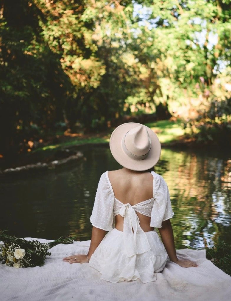 Woman in white backless dress and tan hat sits by a serene tree-lined river with white flowers nearby.