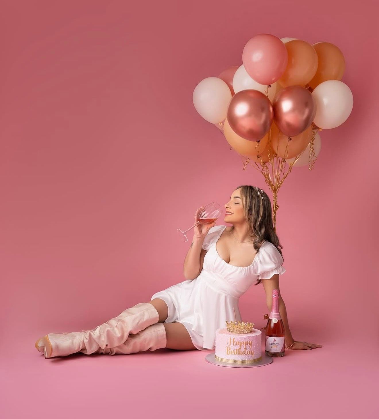 Young woman in white dress and pink boots sipping champagne beside birthday cake with pink and gold balloons.