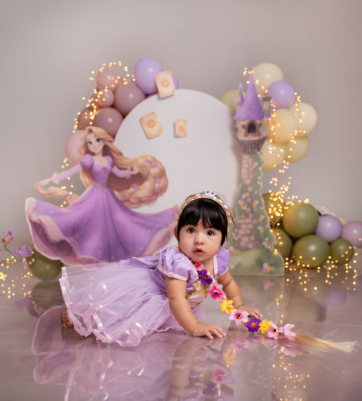 Toddler girl in Rapunzel purple dress poses at a Tangled-themed first birthday party with balloons and fairy lights.