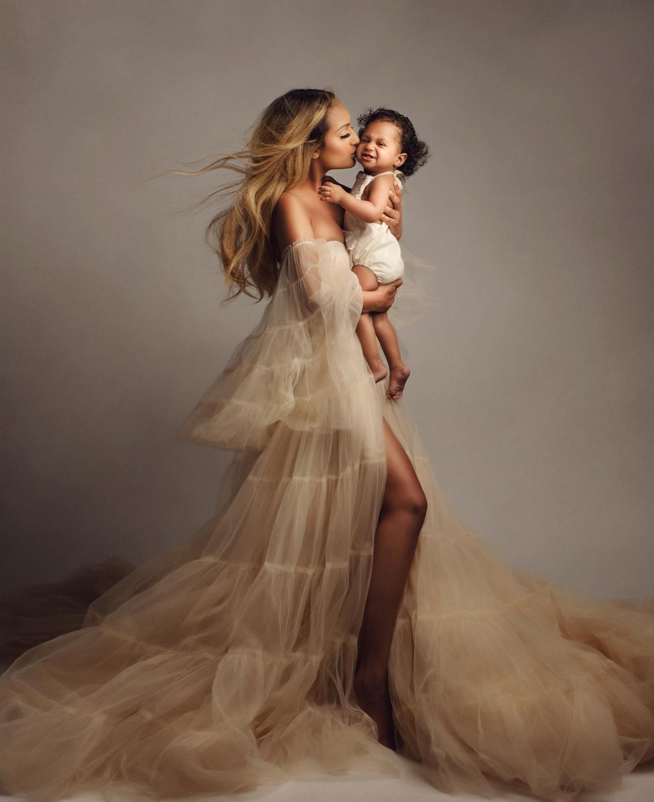 Woman in elegant champagne tulle gown kissing a baby during a professional maternity portrait session.