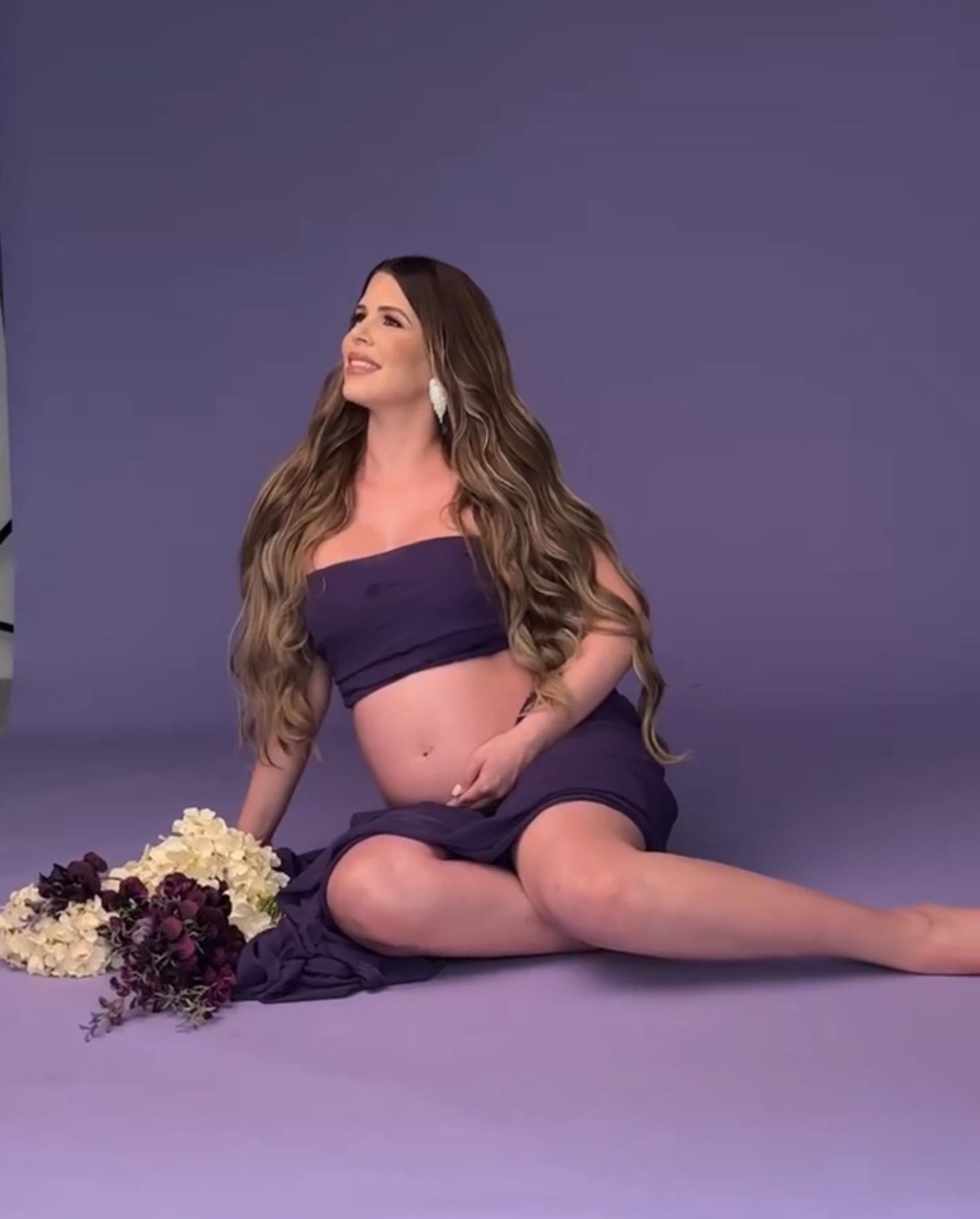 Pregnant woman in purple crop top and skirt sits on floor with flowers during maternity photo shoot.
