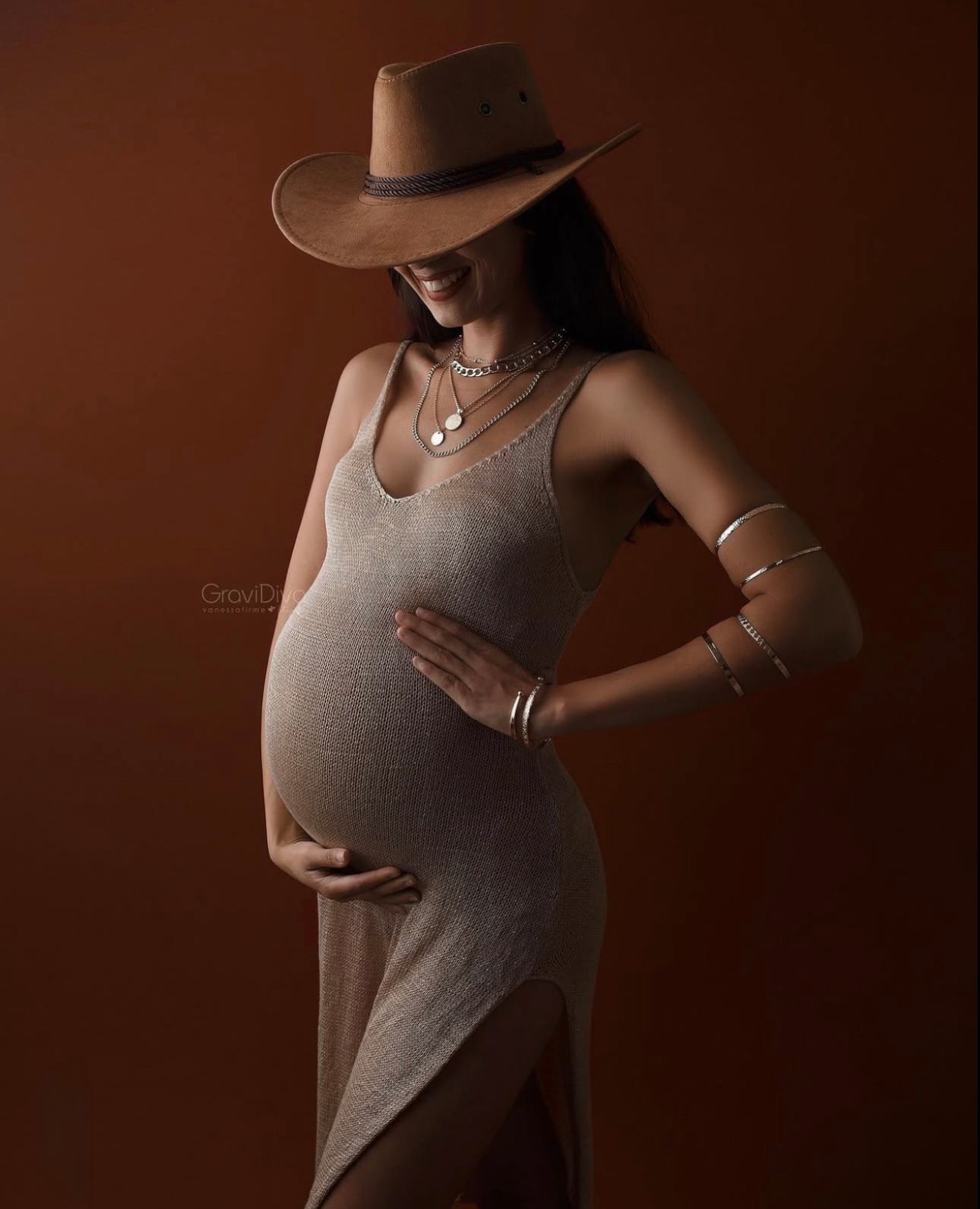 Pregnant woman in gray dress and brown cowboy hat cradling baby bump, wearing silver jewelry on brown background.